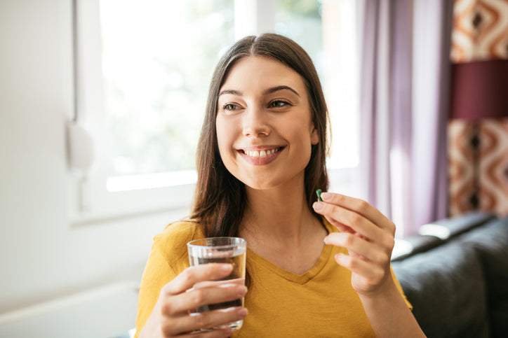 Mulher sorridente segurando um copo de água e um suplemento de magnésio dimalato em ambiente interno, transmitindo saúde e bem-estar.