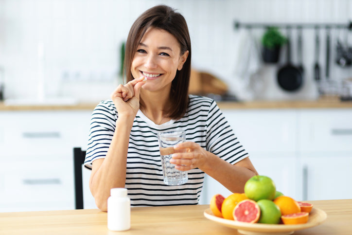 Mulher sorridente segurando um copo de água e uma cápsula de magnésio bisglicinato, com frutas frescas sobre a mesa da cozinha, refletindo hábitos saudáveis e bem-estar.